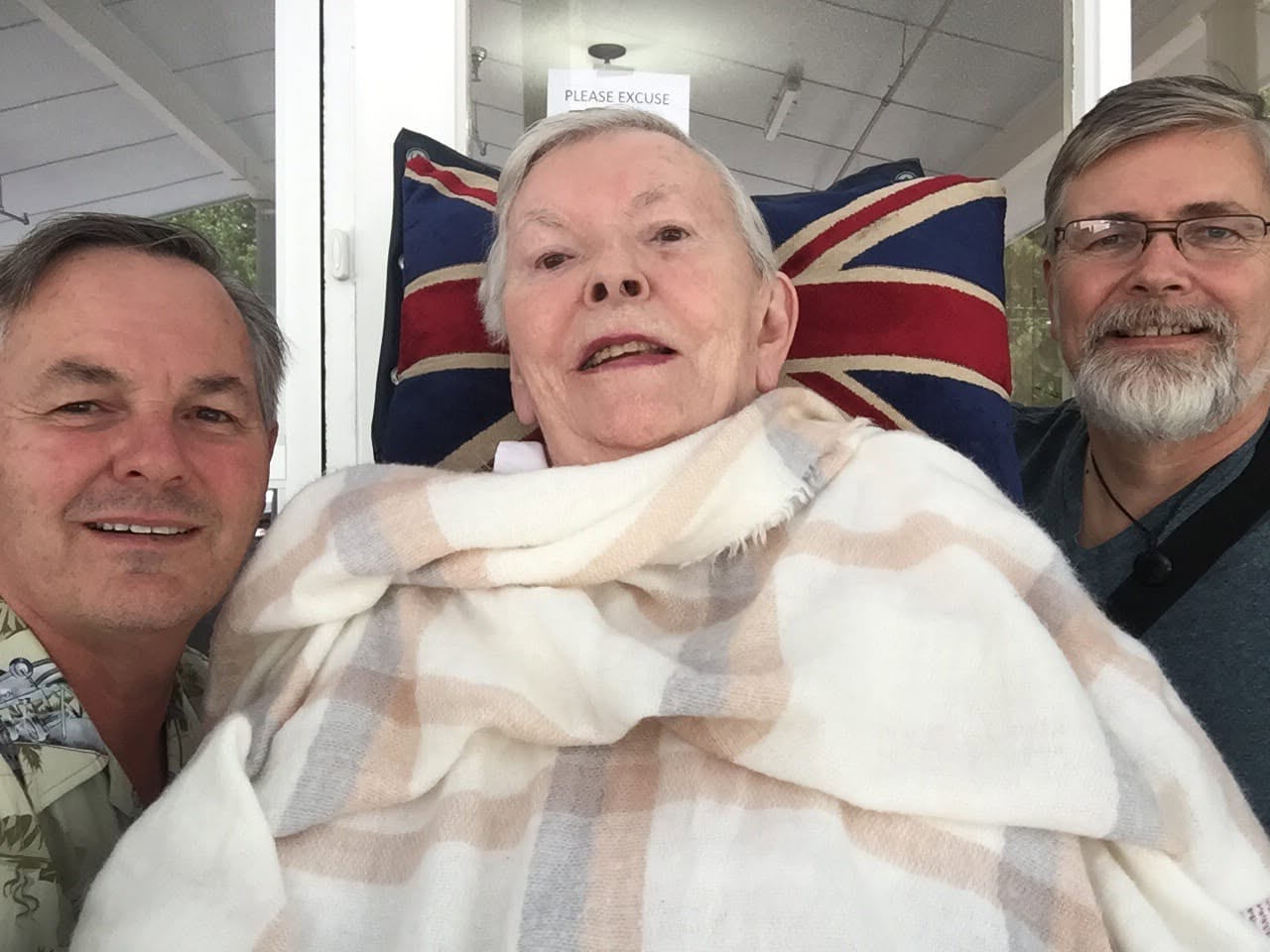 My mother on a covered porch wrapped in a cream and pink checked blanket, a Union Jack cushion propped behind her, and a man leaning in close on either side, all three of us smiling at the camera.