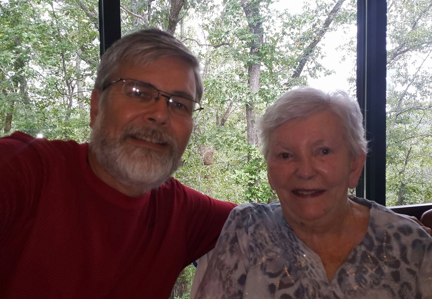My mother and me sitting shoulder to shoulder in front of a tall window, green trees filling the glass behind us.