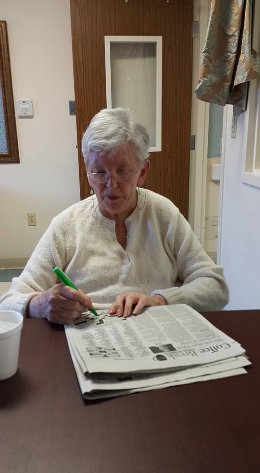 My mother at a table with a green pen, filling in a newspaper crossword and looking down at the page.