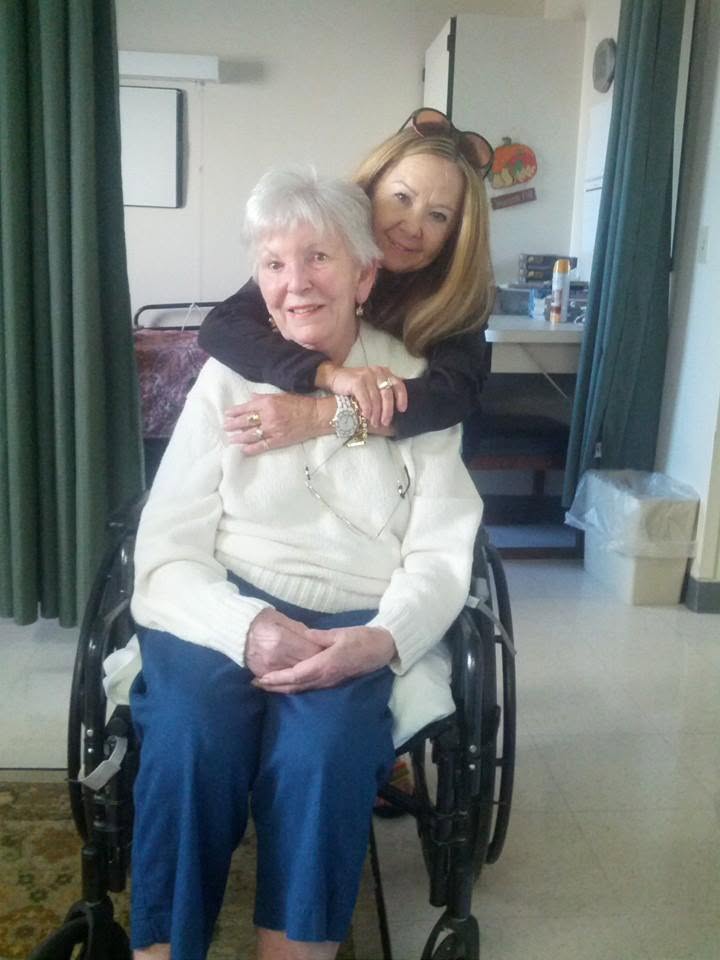 A woman with long fair hair leaning in from behind my mother&rsquo;s shoulder with both arms around her, the two of them smiling together in a plain room.