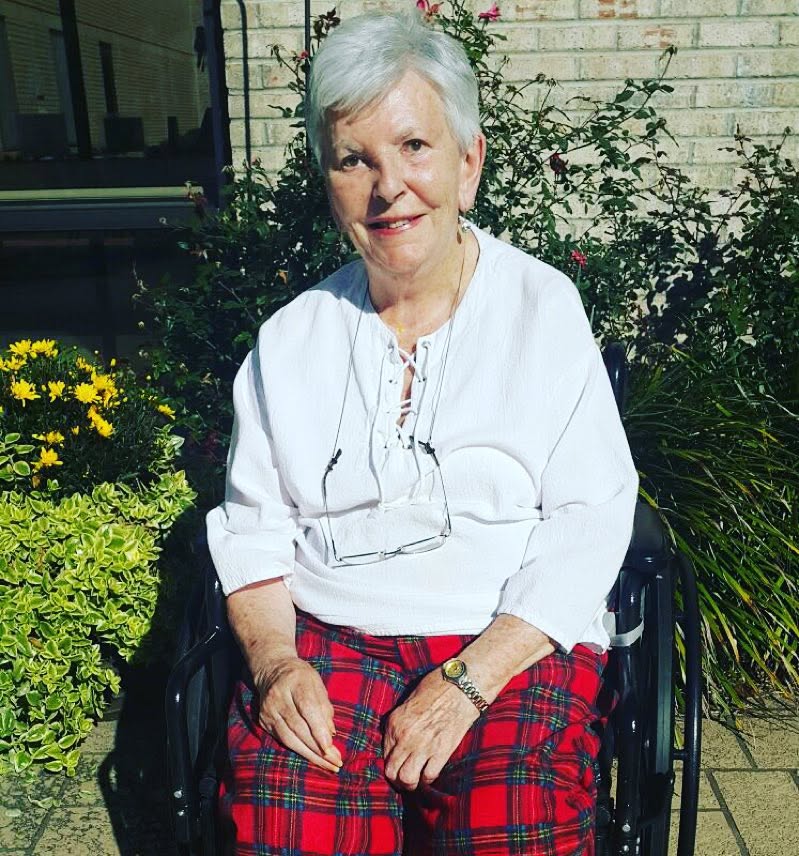 My mother outdoors in bright sunshine in her wheelchair, a yellow flowering shrub beside her and a rose bush against a brick wall behind.