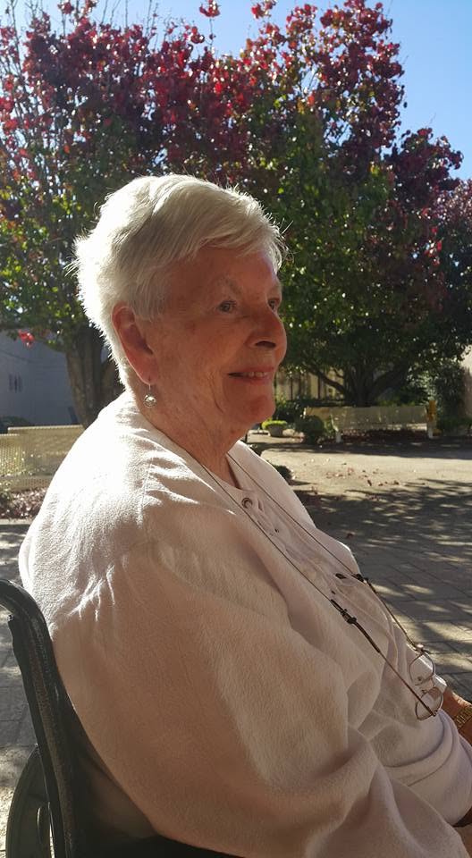 My mother in profile outdoors in low sun, looking away to one side, with a tree in deep red autumn leaf and blue sky behind her.