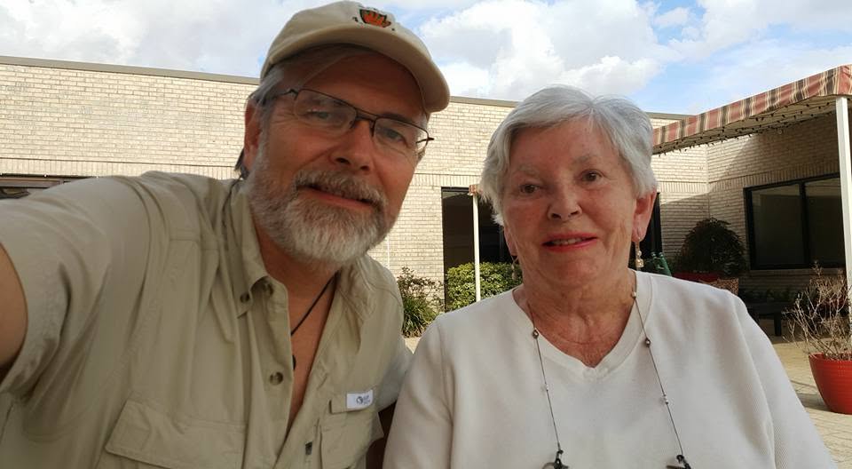 A close arm&rsquo;s-length photograph of my mother and me outdoors in a courtyard, both of us smiling, a brick building and a cloudy sky behind.