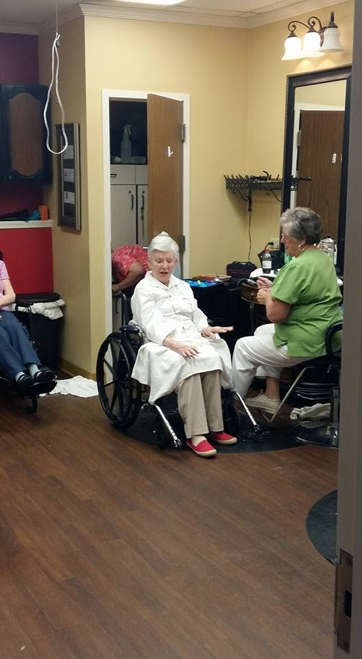 My mother in her wheelchair across a dim salon room with a stylist in green scrubs sitting beside her, photographed from the doorway, with furniture and floor filling the front of the frame.